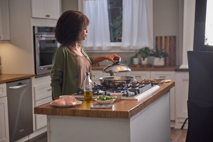 A woman lifting the lid over an A by AMC stainless steel pan on the stovetop.