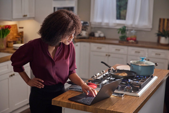 A woman on a laptop at the kitchen island beside a non-stick pan and enamel pot on the stove.