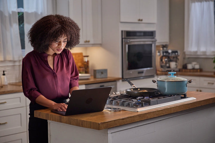 A woman on a laptop at the kitchen island beside an everyday non-stick pan and enamel pot on the stove.